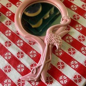 May include: A round hand mirror with a glossy pink frame featuring a sculpted flamingo figure integrated into the handle and side. The flamingo has detailed feather textures and long legs that extend down the length of the handle.