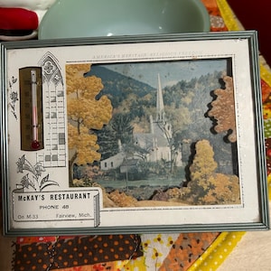 May include: Vintage framed artwork featuring a church scene with fall foliage. The frame includes a thermometer and the text "MCKAY'S RESTAURANT, PHONE 48, Fairview, Mich." The artwork is set against a colorful background.