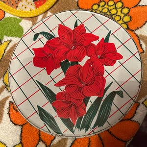 May include: Round metal tray with a white background and a red, green, and black floral design. The tray features a grid pattern and a border. The tray is surrounded by a floral patterned fabric.