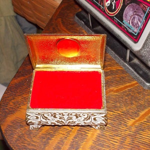 May include: A small, ornate, gold-colored box with a red velvet interior. The box has a hinged lid and intricate floral designs on the sides.