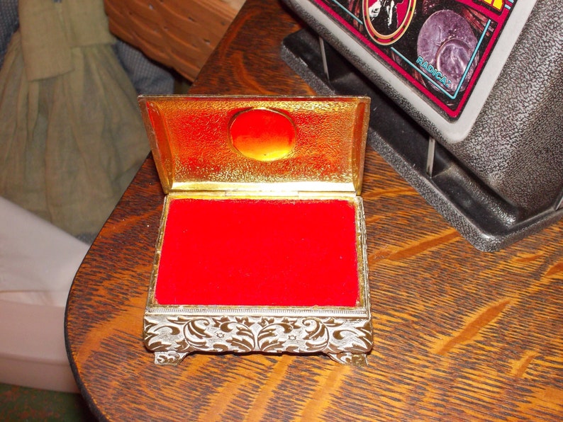 May include: A small, ornate, gold-colored box with a red velvet interior. The box has a hinged lid and intricate floral designs on the sides.