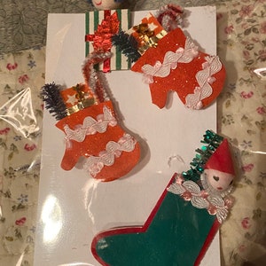May include: A set of three vintage Christmas ornaments featuring a stocking, a mitten, and a doll. The stocking is green with a red trim and the mitten is orange with white lace. The doll is wearing a red hat and has a white face.