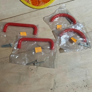 May include: Four red cabinet pulls, each individually packaged in a clear plastic bag. Two of the pulls are U-shaped, and two are arched. Each package includes two silver metal mounting screws. Small yellow price stickers are attached to the bags.