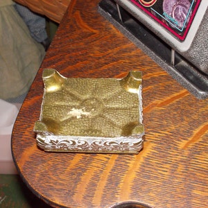 May include: A gold-colored metal trinket box with a decorative white floral design. The box is sitting on a wooden surface.