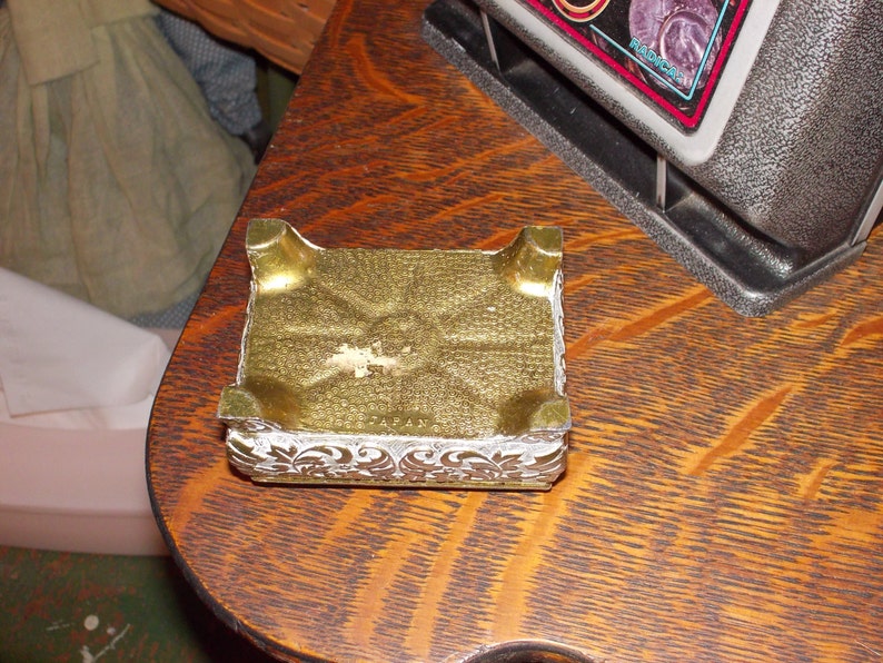 May include: A gold-colored metal trinket box with a decorative white floral design. The box is sitting on a wooden surface.