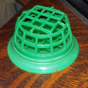 May include: A green plastic floral frog, also known as a flower holder, with a grid-like top and a circular base. The flower arranging tool is designed to hold flower stems in place for floral arrangements.