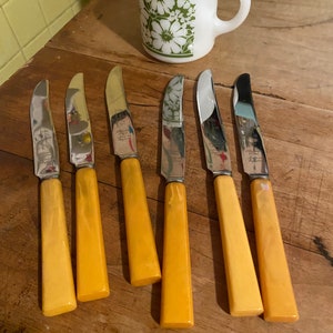 May include: Set of six yellow-handled butter knives with silver blades. The knives are laid out on a wooden surface.