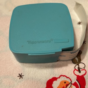 May include: A blue plastic container with a white label attached. The label has the words "Tupperware" and "Contents:" printed on it. The container is sitting on a white tablecloth with a red and white Santa Claus design.