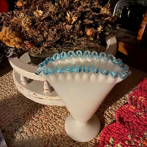May include: A white milk glass vase with a blue scalloped rim. The vase is on a floral patterned surface.