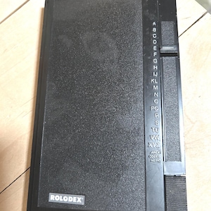 May include: A black Rolodex address book with a textured surface. The book has a vertical letter index on the right side and the Rolodex logo on the bottom left.
