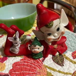May include: A collection of vintage ceramic figurines featuring elves and a small bunny. The elves wear red and green outfits with pointed hats. The figurines are set on a floral patterned quilt, with a green bowl and snack container in the background.