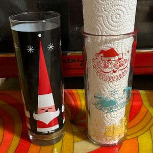 May include: Two vintage holiday-themed drinking glasses. One features a Santa Claus design on a black background with white snowflakes. The other glass holds a roll of paper towels and has a Santa and sleigh design.