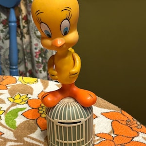 May include: A yellow Tweety Bird figurine standing on top of a gray birdcage. The figurine has orange feet and a small black hair on top of its head. The birdcage has a slot for coins. The background is a floral pattern.