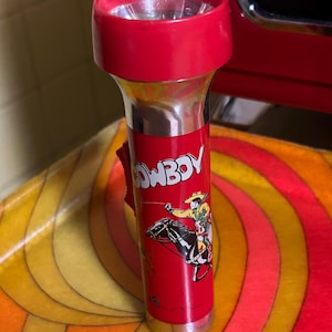 May include: A vintage red flashlight with a silver body. The flashlight features a cowboy graphic and the word "COWBOY" in white. The top and base are red. The flashlight is on a colorful, patterned surface.