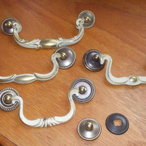 May include: Set of six antique-style cabinet pulls with a white finish and brass accents. The pulls have a decorative, ornate design.