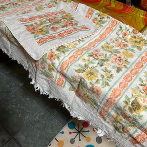May include: White vintage floral towels with a fringed edge. The towels feature a repeating pattern of peach, yellow, and green flowers with orange and green accents. A matching square hand towel is also visible.