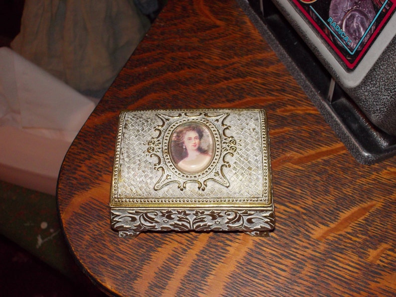 May include: A small, ornate, silver-colored trinket box with a white and gold design. The box has a raised, oval-shaped image of a woman with dark hair and a white dress. The box is sitting on a wooden surface.
