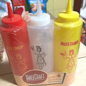 May include: A set of three squeeze dispenser bottles for condiments. The red bottle is labeled "KETCHUP", the clear bottle is unlabeled, and the yellow bottle is labeled "MUSTARD". Each bottle has a matching illustration of a waitress.