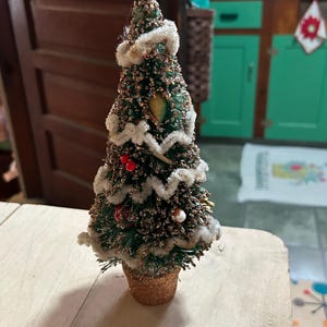 May include: A miniature artificial Christmas tree with dark green bristles, gold glitter accents, and white pipe cleaner garland. The tree is decorated with small red berries and sits in a textured, glittery gold-colored pot base.