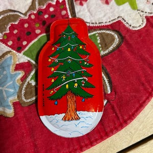 May include: A red, Christmas-themed metal tray shaped like a bottle, featuring a decorated green Christmas tree with ornaments and a brown trunk. The tray is set on a red and white Christmas tree skirt. The text "MADE IN JAPAN" is visible.