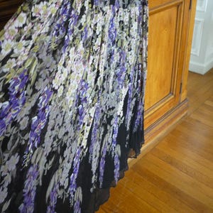 Puede incluir: Una falda larga con un estampado floral en tonos morados, blancos y verdes sobre un fondo negro. La falda parece estar hecha de una tela ligera, con un dise&ntilde;o plisado. La falda cuelga cerca de un mueble de madera y un suelo de parquet.
