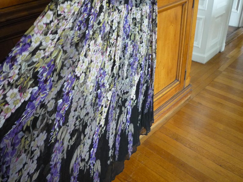 Puede incluir: Una falda larga con un estampado floral en tonos morados, blancos y verdes sobre un fondo negro. La falda parece estar hecha de una tela ligera, con un dise&ntilde;o plisado. La falda cuelga cerca de un mueble de madera y un suelo de parquet.