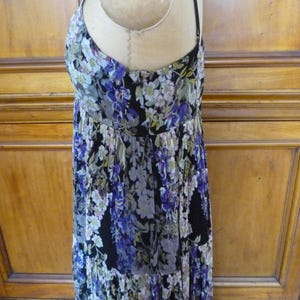 Puede incluir: Vestido con estampado floral sobre fondo negro, con una mezcla de flores moradas, blancas y verdes. El vestido tiene tirantes finos y una falda fluida con volantes. La tela parece ligera, adecuada para climas c&aacute;lidos.