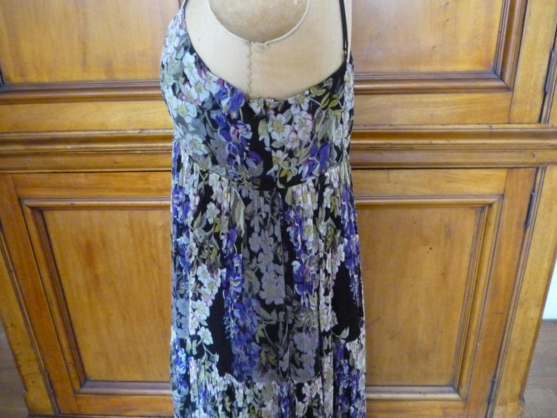 Puede incluir: Vestido con estampado floral sobre fondo negro, con una mezcla de flores moradas, blancas y verdes. El vestido tiene tirantes finos y una falda fluida con volantes. La tela parece ligera, adecuada para climas c&aacute;lidos.