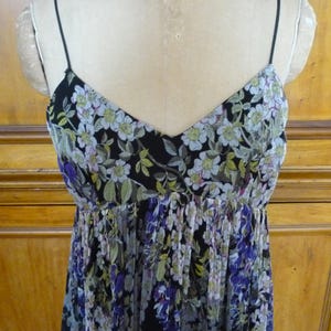 Puede incluir: Vestido con estampado floral con escote en V y tirantes finos. El vestido presenta un fondo negro con un estampado de flores blancas, verdes y moradas. El corpi&ntilde;o es entallado y la falda fluida.