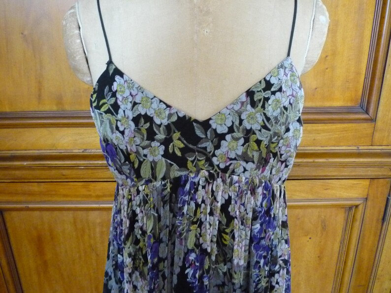 Puede incluir: Vestido con estampado floral con escote en V y tirantes finos. El vestido presenta un fondo negro con un estampado de flores blancas, verdes y moradas. El corpi&ntilde;o es entallado y la falda fluida.