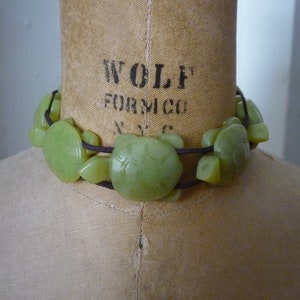 May include: A green jade turtle necklace with brown cord. The necklace is on a fabric-covered mannequin bust.