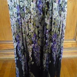 Puede incluir: Vestido largo con estampado floral. El vestido presenta una base negra con dise&ntilde;os florales blancos, morados y verdes en cascada. La tela parece ligera y adecuada para climas c&aacute;lidos.