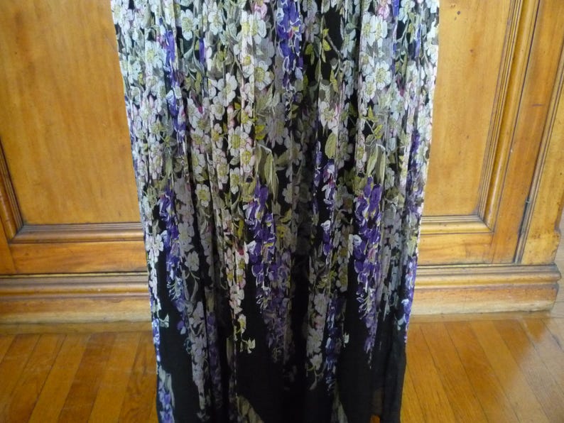 Puede incluir: Vestido largo con estampado floral. El vestido presenta una base negra con dise&ntilde;os florales blancos, morados y verdes en cascada. La tela parece ligera y adecuada para climas c&aacute;lidos.