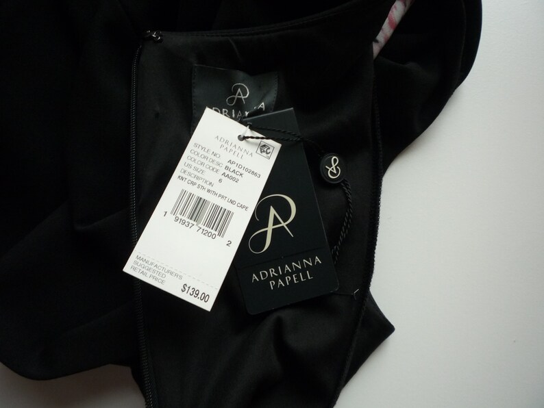 May include: Black knit dress with a matching knit cape. The dress has a zipper closure and a tag that reads "Adrianna Papell" with the style number API0102863, color code AA002, and size 6. The suggested retail price is $139.00.