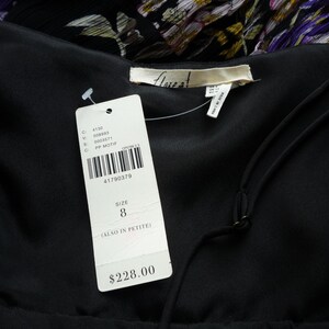 Puede incluir: Primer plano de un vestido negro con una etiqueta adjunta. La etiqueta muestra un c&oacute;digo de barras, la talla 8 y el nombre de la marca. El vestido tiene cuello en V y tirantes finos. El fondo muestra un estampado floral.