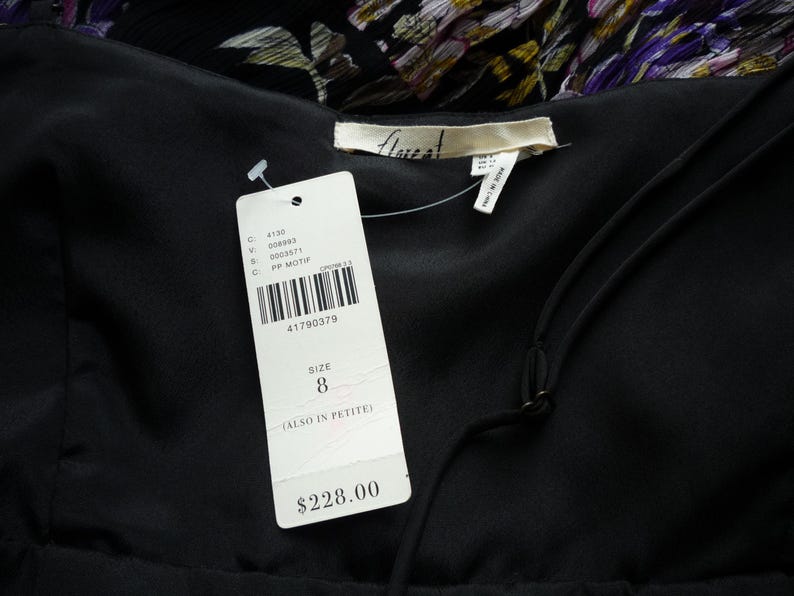 Puede incluir: Primer plano de un vestido negro con una etiqueta adjunta. La etiqueta muestra un c&oacute;digo de barras, la talla 8 y el nombre de la marca. El vestido tiene cuello en V y tirantes finos. El fondo muestra un estampado floral.