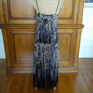 Puede incluir: Un vestido largo con estampado floral y tirantes finos. El vestido presenta un fondo negro con un estampado de flores moradas, blancas y verdes. El vestido se exhibe sobre un maniqu&iacute; contra un fondo de madera.