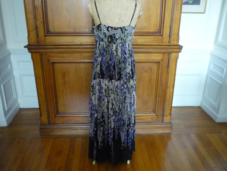 Puede incluir: Un vestido largo con estampado floral y tirantes finos. El vestido presenta un fondo negro con un estampado de flores moradas, blancas y verdes. El vestido se exhibe sobre un maniqu&iacute; contra un fondo de madera.