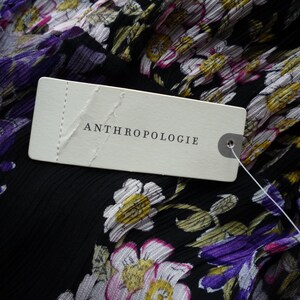 Puede incluir: Primer plano de una tela negra con un estampado floral en morado, blanco y amarillo. Una etiqueta blanca con la palabra "ANTHROPOLOGIE" est&aacute; adherida a la tela.