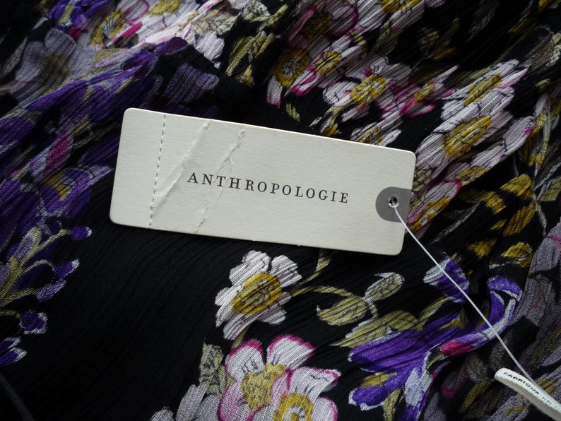 Puede incluir: Primer plano de una tela negra con un estampado floral en morado, blanco y amarillo. Una etiqueta blanca con la palabra "ANTHROPOLOGIE" est&aacute; adherida a la tela.