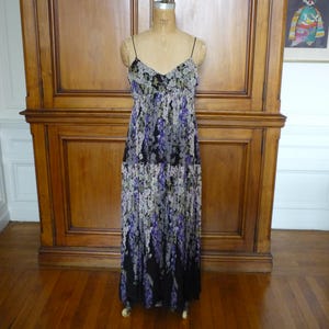 Puede incluir: Vestido largo con tirantes finos y estampado floral, expuesto sobre un maniqu&iacute;. El vestido presenta un escote en V y una falda fluida con un patr&oacute;n de flores moradas, blancas y negras. El vestido est&aacute; sobre un fondo de madera.