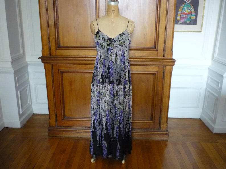 Puede incluir: Vestido largo con tirantes finos y estampado floral, expuesto sobre un maniqu&iacute;. El vestido presenta un escote en V y una falda fluida con un patr&oacute;n de flores moradas, blancas y negras. El vestido est&aacute; sobre un fondo de madera.