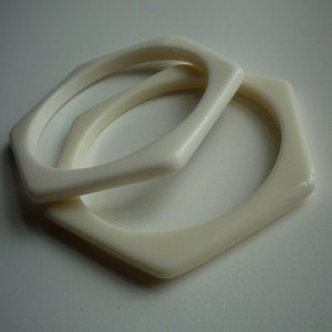 May include: Two white plastic bangles, one slightly larger than the other, are stacked on top of each other. The bangles are both oval shaped with rounded edges.