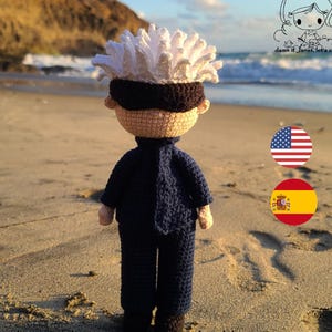 Op de afbeelding: Een gehaakt figuur met wit haar, een zwart oogmasker en een blauw pak staat op een zandstrand. Het figuur staat met de rug naar de camera. De Amerikaanse en Spaanse vlag zijn zichtbaar op de achtergrond.