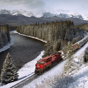 May include: A red train engine with the text "Canadian Pacific" on the side is pulling a long train of boxcars along a snowy track through a wintery forest. The train is traveling along a river with snow-covered mountains in the background.