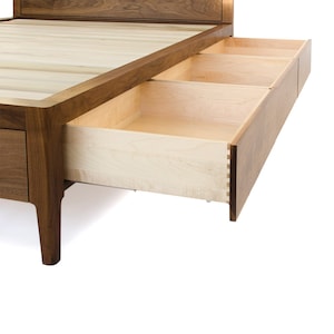 Peut inclure: Un cadre de lit en bois avec une t&ecirc;te de lit en bois clair et une base en bois fonc&eacute;. Le cadre de lit a trois tiroirs qui sont partiellement ouverts, r&eacute;v&eacute;lant un int&eacute;rieur en bois clair.