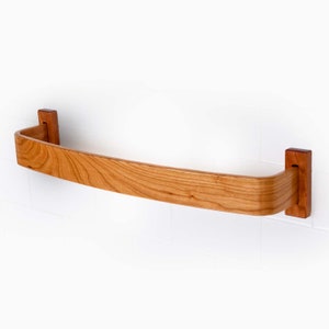 May include: A curved wooden towel rack with a natural cherry finish. The rack is mounted to a white tiled wall with two wooden brackets.