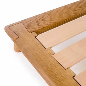May include: Close-up of a wooden bed frame with a slatted base. The frame is made of light brown wood with a natural finish. The slats are made of a lighter wood and are arranged in a grid pattern.