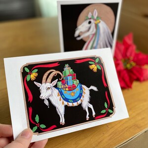 May include: A white goat with a red, blue, and green saddle carrying a stack of wrapped gifts. The goat is standing on a black background with red, green, and gold accents. The image is framed with a red ribbon and holly.