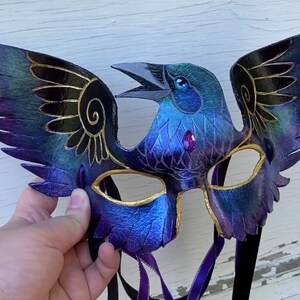 May include: A purple and blue leather masquerade mask with gold accents and a bird design. The mask has two wings and a bird's head with a jeweled eye.